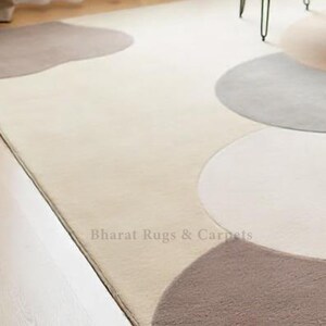 May include: A cream-colored rug with a modern design featuring overlapping circles in shades of gray and brown. The rug is made of soft, plush material and has a subtle sheen. The text "Bharat Rugs & Carpets" is visible in the lower right corner of the rug.