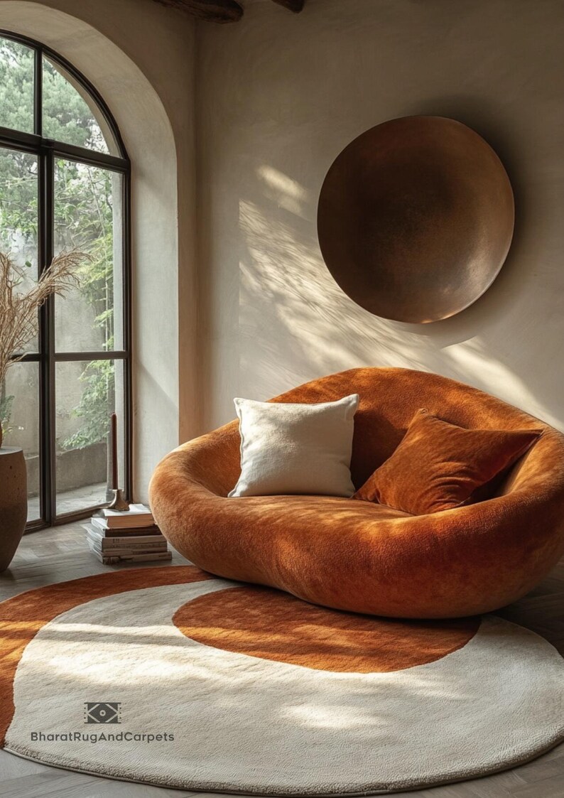 May include: A round, cream-colored rug with a brown, curved design. A large, orange, plush sofa with two pillows sits on the rug. A large, round, copper-colored wall decor hangs on the wall behind the sofa.