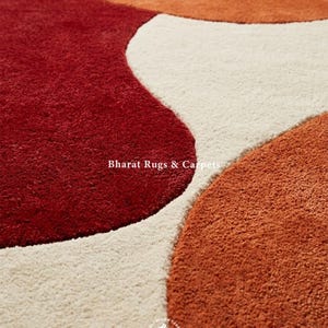 May include: A plush area rug featuring a pattern of large, organic, rounded shapes in deep red, burnt orange, and cream. The text Bharat Rugs & Carpets is printed in white across the center, with a circular logo at the bottom containing the same text.