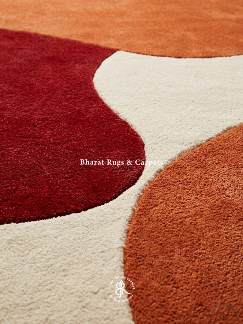 May include: A plush area rug featuring a pattern of large, organic, rounded shapes in deep red, burnt orange, and cream. The text Bharat Rugs & Carpets is printed in white across the center, with a circular logo at the bottom containing the same text.