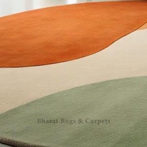 Puede incluir: Una alfombra redonda abstracta en tres colores: naranja, beige y verde. La alfombra tiene una forma ondulada org&aacute;nica y una textura sutil. El texto "Bharat Rugs & Carpets" est&aacute; impreso en la alfombra.