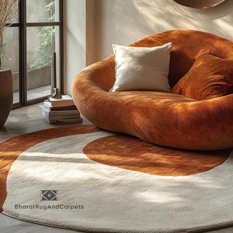 May include: A round, cream-colored rug with two brown, semi-circular patterns. The rug is in front of a brown, plush, modern sofa with white pillows. The text "BharatRugAndCarpets" is visible in the bottom left corner of the rug.