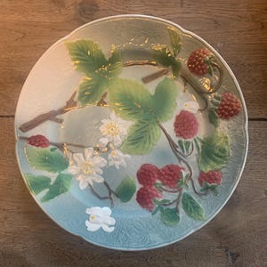 Può includere: Un piatto rotondo in ceramica con un disegno in rilievo di lamponi, foglie e fiori. Il piatto ha uno sfondo azzurro e bianco con foglie verdi e lamponi rossi. Il piatto è probabilmente per uso decorativo.