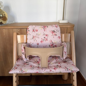 May include: A wooden high chair featuring a light pink fabric cushion set with a floral pattern of red roses and green leaves. The chair includes a beige plastic safety guard and backrest support, with a natural wood frame and a black footrest bar.