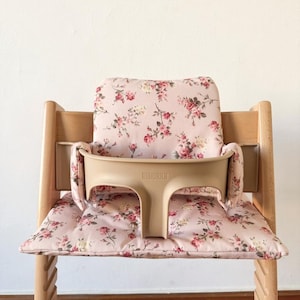 May include: A light-colored wooden high chair featuring a beige plastic safety rail with the embossed text STOKKE. The chair includes a soft, padded seat and backrest cushion covered in a light pink fabric with a floral pattern of red roses.