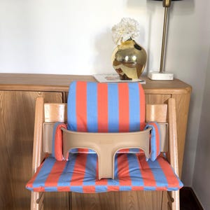 May include: A wooden high chair featuring a padded seat and backrest cushion with a vertical orange and light blue striped pattern. The chair includes a beige plastic safety guard with side supports that attach to the frame for secure seating.