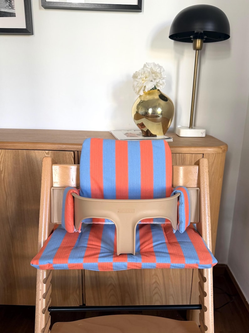 May include: A wooden high chair featuring a padded seat and backrest cushion with a vertical orange and light blue striped pattern. The chair includes a beige plastic safety guard with side supports that attach to the frame for secure seating.