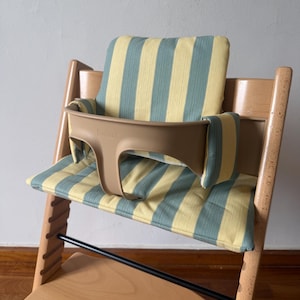Op de afbeelding: Een houten kinderstoel met een lichtbruin kunststof babyzitje waarop de merknaam Stokke aan de voorzijde is gedrukt. De stoel is voorzien van een gewatteerd zit- en rugkussen met een verticaal gestreept patroon in lichtgeel en saliegroen.