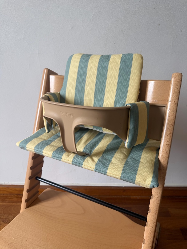 Op de afbeelding: Een houten kinderstoel met een lichtbruin kunststof babyzitje waarop de merknaam Stokke aan de voorzijde is gedrukt. De stoel is voorzien van een gewatteerd zit- en rugkussen met een verticaal gestreept patroon in lichtgeel en saliegroen.