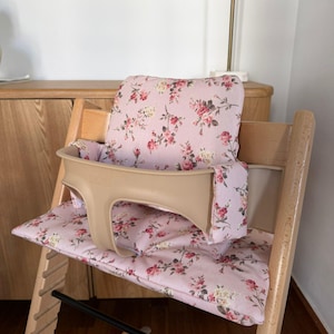May include: A wooden high chair featuring a light pink fabric cushion set with a floral pattern of red roses and green leaves. The chair includes a beige plastic safety guard that wraps around the front and sides of the seat area.