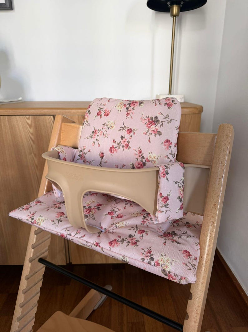 May include: A wooden high chair featuring a light pink fabric cushion set with a floral pattern of red roses and green leaves. The chair includes a beige plastic safety guard that wraps around the front and sides of the seat area.