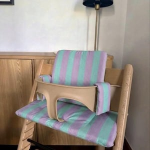 May include: A wooden high chair featuring a light beige plastic safety guard and a padded seat cushion with a vertical striped pattern in alternating shades of soft lavender and mint green, covering both the backrest and the seat base.