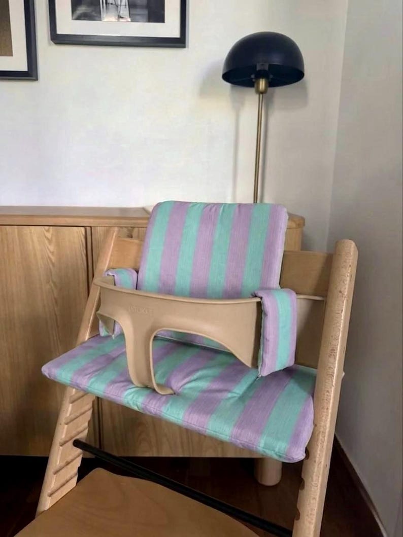 May include: A wooden high chair featuring a light beige plastic safety guard and a padded seat cushion with a vertical striped pattern in alternating shades of soft lavender and mint green, covering both the backrest and the seat base.