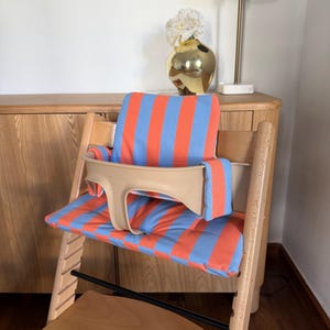 May include: A wooden high chair featuring a light blue and orange striped fabric cushion set. The cushion set includes a backrest pad and a seat pad, complemented by a beige plastic safety rail that secures around the seat area for added support.
