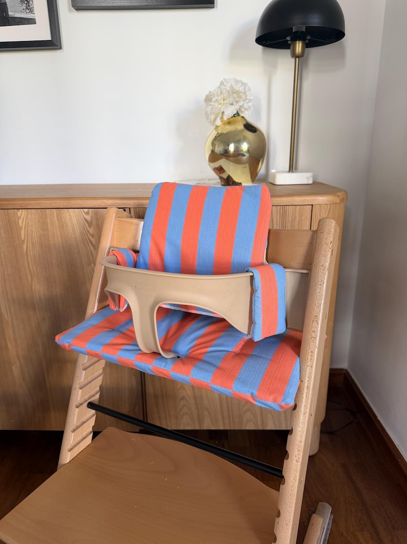 May include: A wooden high chair featuring a light blue and orange striped fabric cushion set. The cushion set includes a backrest pad and a seat pad, complemented by a beige plastic safety rail that secures around the seat area for added support.