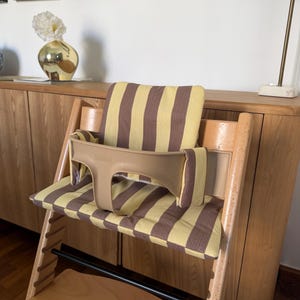May include: A wooden high chair featuring a light brown plastic safety rail and a padded seat cushion with a vertical yellow and brown striped pattern. The chair has a natural wood frame with adjustable slots and a flat wooden footrest at the base.
