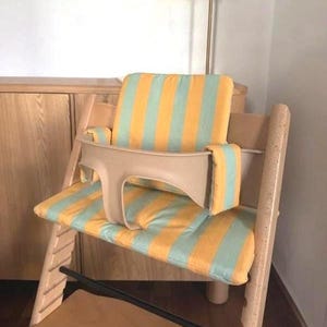 May include: A wooden high chair with a beige safety bar and a striped cushion in yellow and light blue. The chair is designed for a child and is placed in a room with a lamp and wooden furniture.