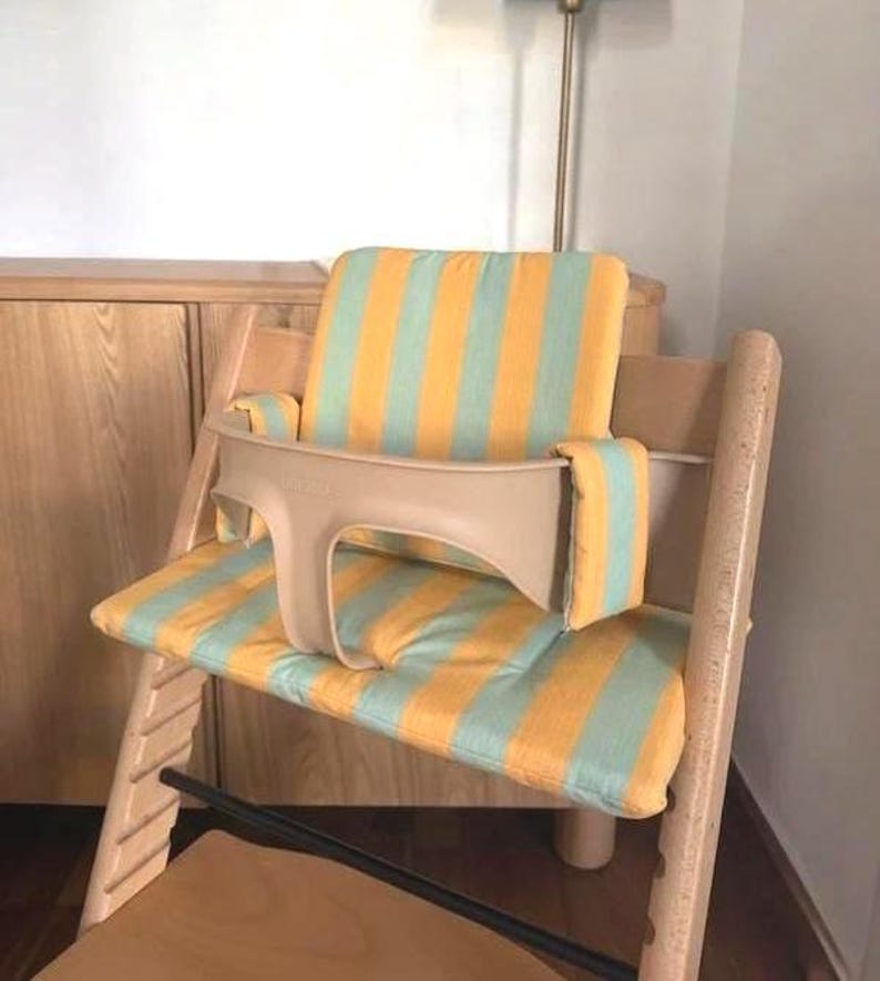 May include: A wooden high chair with a beige safety bar and a striped cushion in yellow and light blue. The chair is designed for a child and is placed in a room with a lamp and wooden furniture.