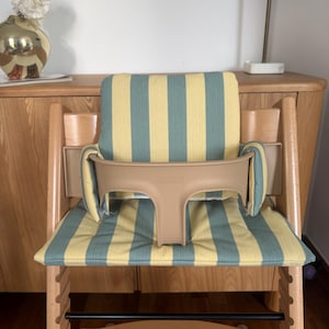 Op de afbeelding: Een houten kinderstoel met een lichtbeige kunststof veiligheidsbeugel en een gewatteerd zitkussen met een verticaal streeppatroon in gedempt geel en saliegroen. De stoel heeft een natuurlijke houten afwerking en een zwarte voetensteun.