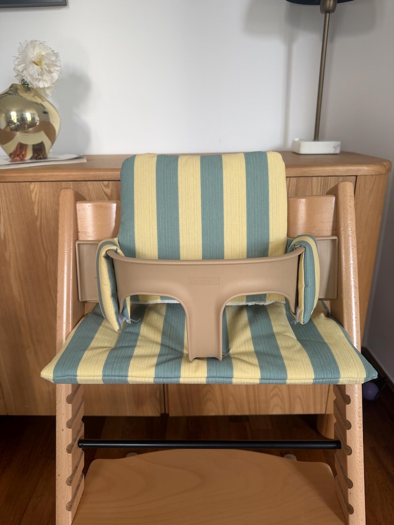 Op de afbeelding: Een houten kinderstoel met een lichtbeige kunststof veiligheidsbeugel en een gewatteerd zitkussen met een verticaal streeppatroon in gedempt geel en saliegroen. De stoel heeft een natuurlijke houten afwerking en een zwarte voetensteun.