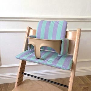 May include: A wooden high chair featuring a light beige plastic safety rail and a padded seat cushion with a vertical striped pattern in alternating shades of mint green and lavender. The chair has a natural wood finish and a footrest.