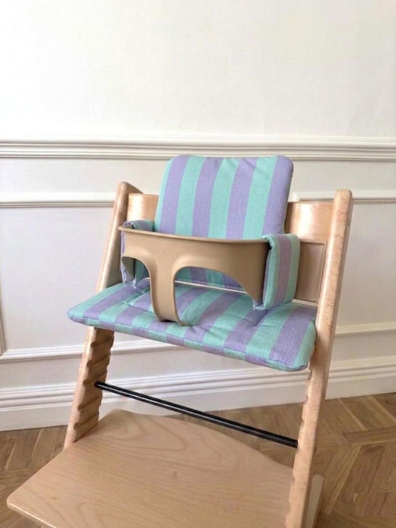 May include: A wooden high chair featuring a light beige plastic safety rail and a padded seat cushion with a vertical striped pattern in alternating shades of mint green and lavender. The chair has a natural wood finish and a footrest.