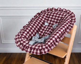 May include: A high chair seat cushion featuring a burgundy and white gingham check pattern with a ruffled edge. The cushion includes light grey safety straps and is mounted on a light-coloured wooden high chair frame with a footrest.