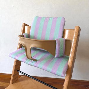 May include: A wooden high chair featuring a light green and lavender striped fabric cushion set. The set includes a backrest pad, a seat pad, and two side arm covers. A tan plastic safety rail is attached to the front of the wooden frame.