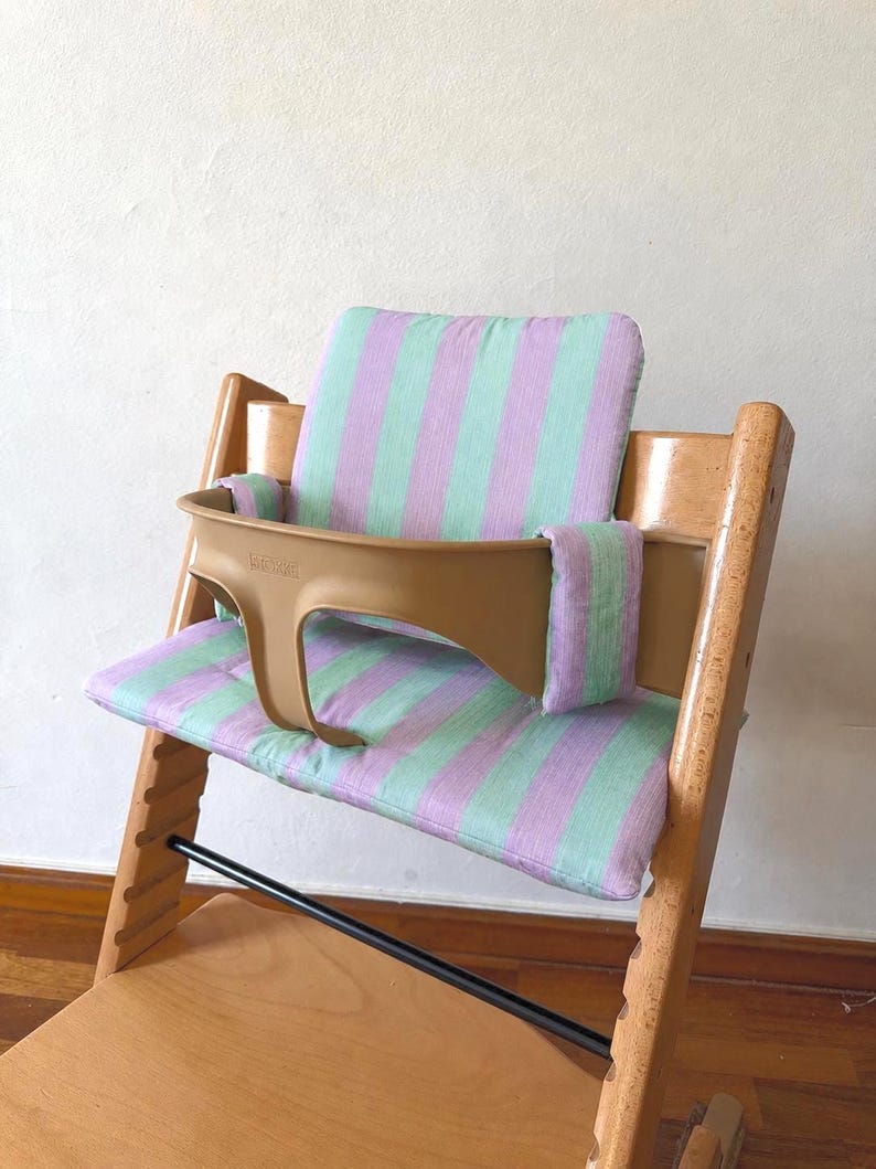 May include: A wooden high chair featuring a light green and lavender striped fabric cushion set. The set includes a backrest pad, a seat pad, and two side arm covers. A tan plastic safety rail is attached to the front of the wooden frame.