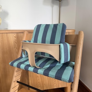 May include: A wooden high chair featuring a beige plastic safety guard and a padded seat cushion with a vertical striped pattern in teal and dark blue. The chair has a light wood frame with a horizontal black metal bar across the front.