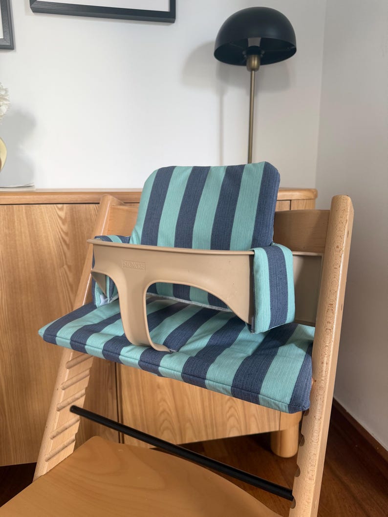 May include: A wooden high chair featuring a beige plastic safety guard and a padded seat cushion with a vertical striped pattern in teal and dark blue. The chair has a light wood frame with a horizontal black metal bar across the front.