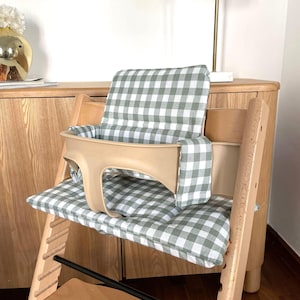 May include: A wooden high chair with a beige safety harness and a green and white checkered cushion. The chair has a wooden frame and is designed for a baby or toddler.