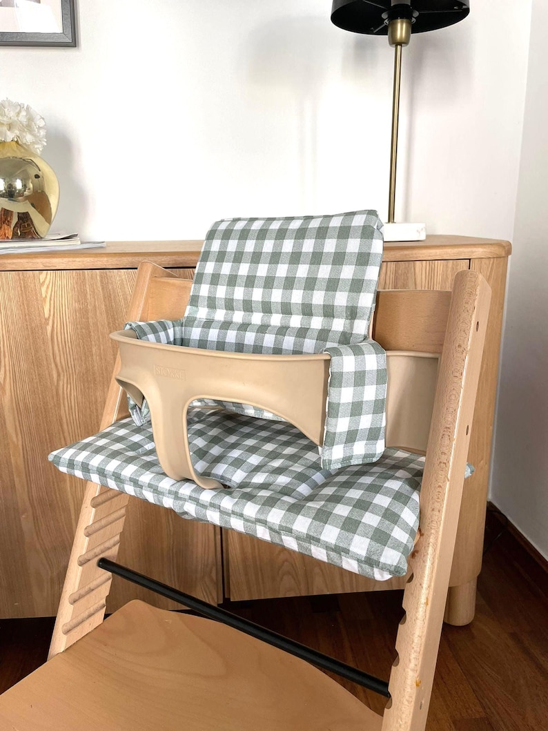 May include: A wooden high chair with a beige safety harness and a green and white checkered cushion. The chair has a wooden frame and is designed for a baby or toddler.