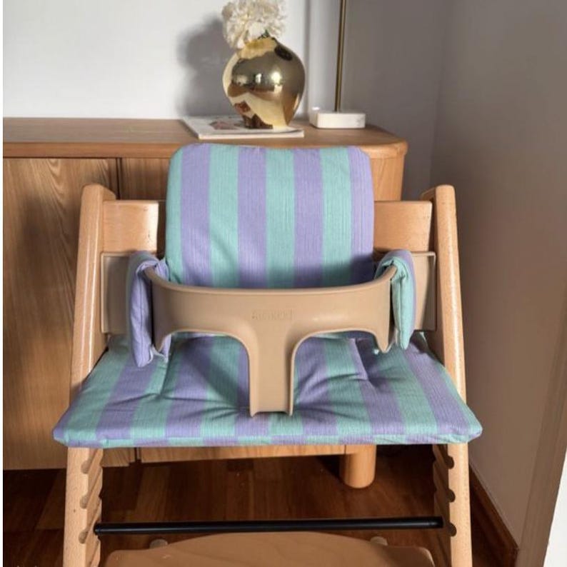 May include: A wooden high chair featuring a light purple and mint green striped fabric cushion set. The chair includes a beige plastic safety rail and a matching backrest pad, providing a structured seat for a child with a natural wood frame.