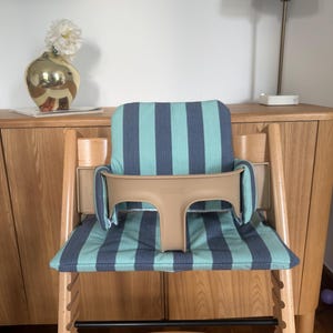 May include: A wooden high chair featuring a light wood frame and a beige plastic safety rail. The seat and backrest are covered in a fabric cushion with vertical stripes in alternating shades of teal and dark blue, providing support.