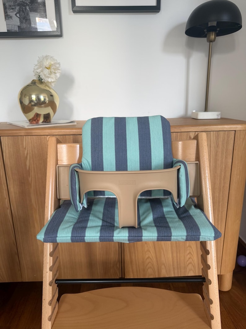 May include: A wooden high chair featuring a light wood frame and a beige plastic safety rail. The seat and backrest are covered in a fabric cushion with vertical stripes in alternating shades of teal and dark blue, providing support.