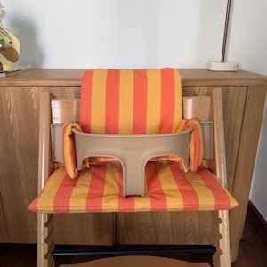 May include: A wooden high chair with a beige safety bar and a seat cushion with orange and yellow stripes. The chair has a footrest and is designed for a child. The chair is made of light-colored wood.