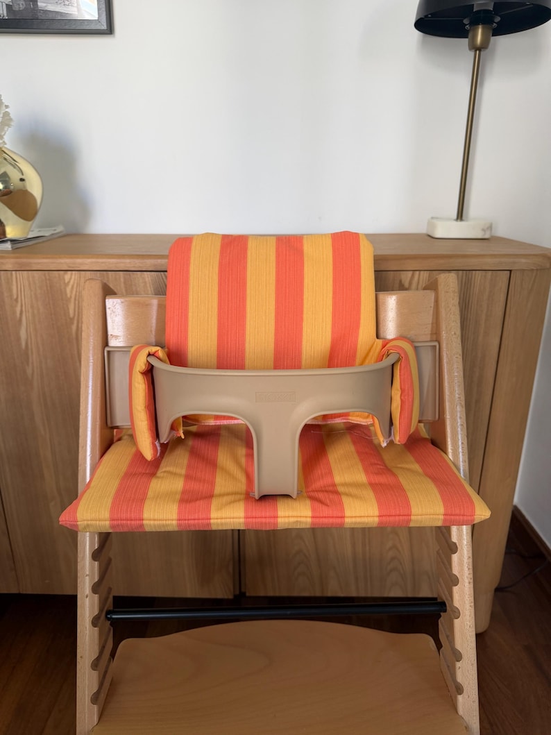May include: A wooden high chair with a beige safety bar and a seat cushion with orange and yellow stripes. The chair has a footrest and is designed for a child. The chair is made of light-colored wood.