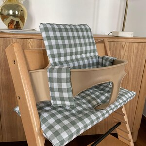 May include: A wooden high chair with a beige seat and backrest, featuring a green and white checkered cushion. The chair has a footrest and is designed for a child. The chair is placed in front of a wooden cabinet.