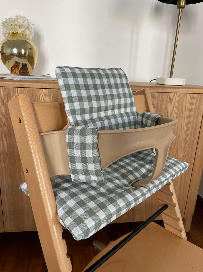 May include: A wooden high chair with a beige seat and backrest, featuring a green and white checkered cushion. The chair has a footrest and is designed for a child. The chair is placed in front of a wooden cabinet.