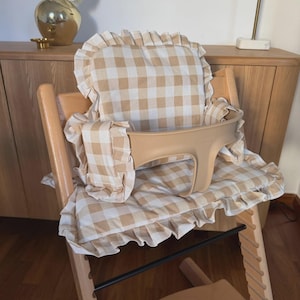 May include: A wooden high chair featuring a beige and white gingham fabric cushion set with ruffled edges. The set includes a backrest cushion and a seat pad, complemented by a smooth, light-toned plastic safety rail attached to the wooden frame.