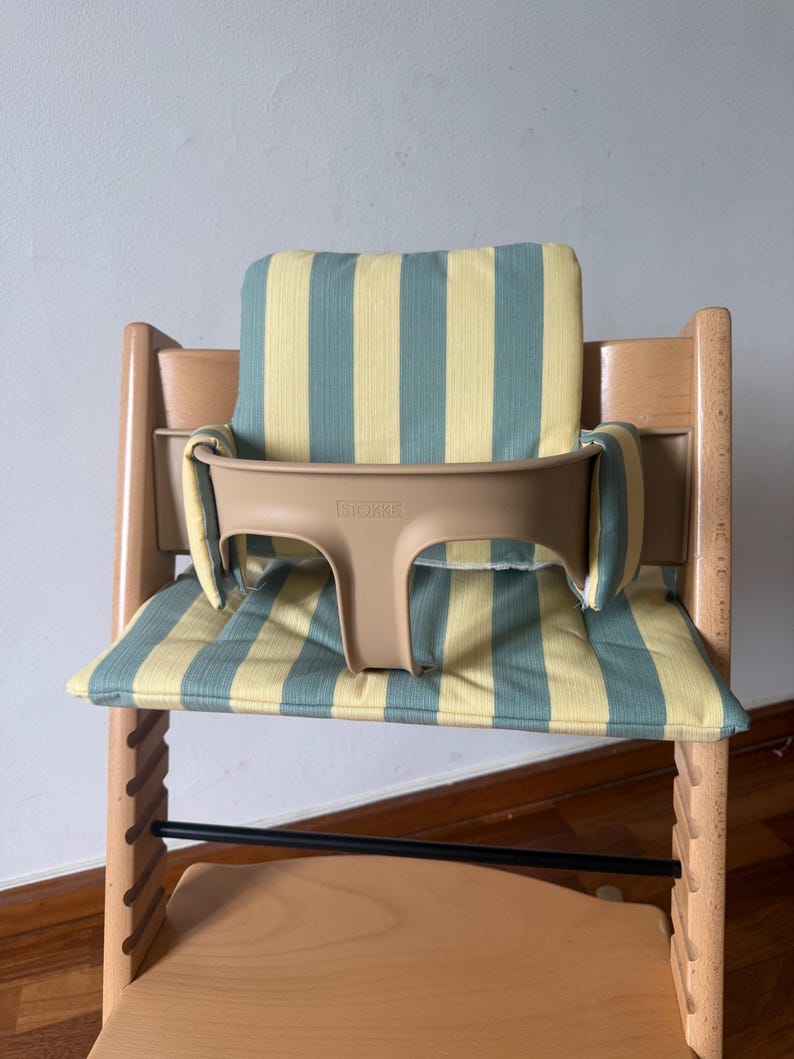 Op de afbeelding: Een houten kinderstoel met een lichtbruine kunststof babyzitje met de merknaam STOKKE in reli&euml;f op de voorzijde. De stoel is voorzien van een gewatteerd zitkussen en rugkussen, beide versierd met een verticaal gestreept patroon in geel en saliegroen.
