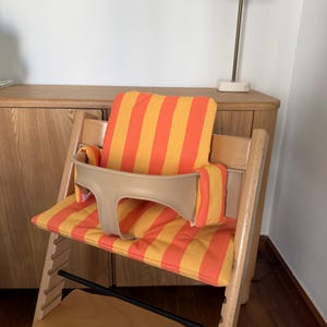 May include: A wooden high chair with a beige safety bar and a striped orange and yellow cushion. The chair is designed for a child and has a footrest. The chair is made of light-colored wood.