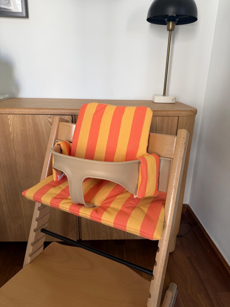 May include: A wooden high chair with a beige safety bar and a striped orange and yellow cushion. The chair is designed for a child and has a footrest. The chair is made of light-colored wood.
