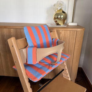 May include: A wooden high chair featuring a light beige plastic safety rail and a padded cushion set with a vibrant orange and light blue vertical striped pattern. The cushion set includes a backrest pad and a seat pad for the high chair.