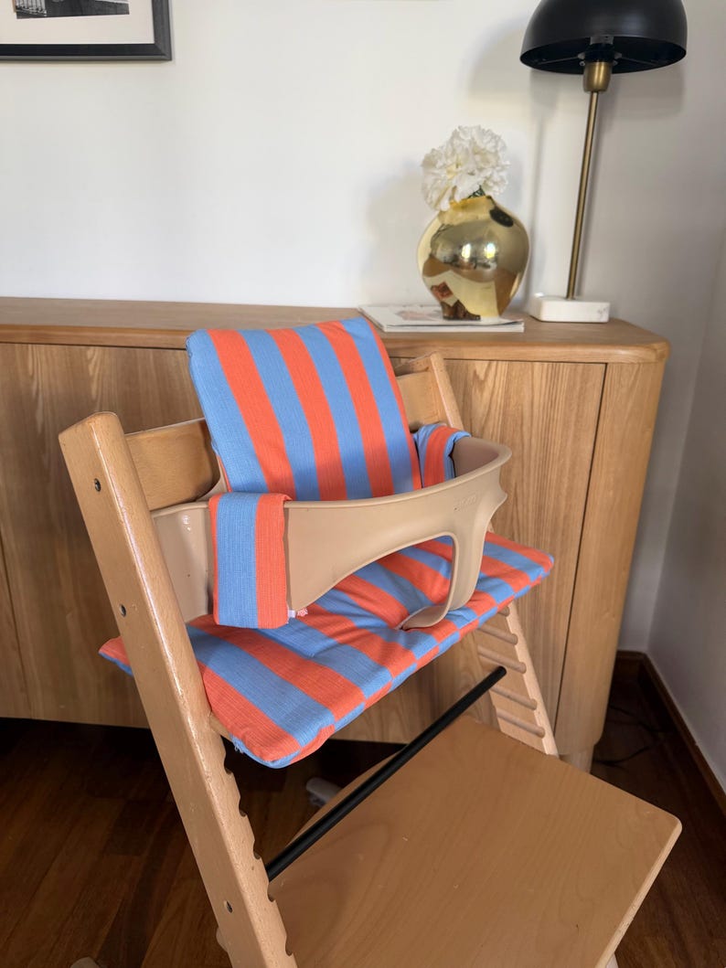 May include: A wooden high chair featuring a light beige plastic safety rail and a padded cushion set with a vibrant orange and light blue vertical striped pattern. The cushion set includes a backrest pad and a seat pad for the high chair.