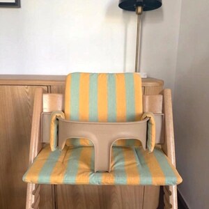 Op de afbeelding: Houten kinderstoel met een beige veiligheidsbeugel en een zitkussen met gele en lichtgroene strepen. De stoel is ontworpen voor een kind en staat in een kamer met een lamp en houten meubels.