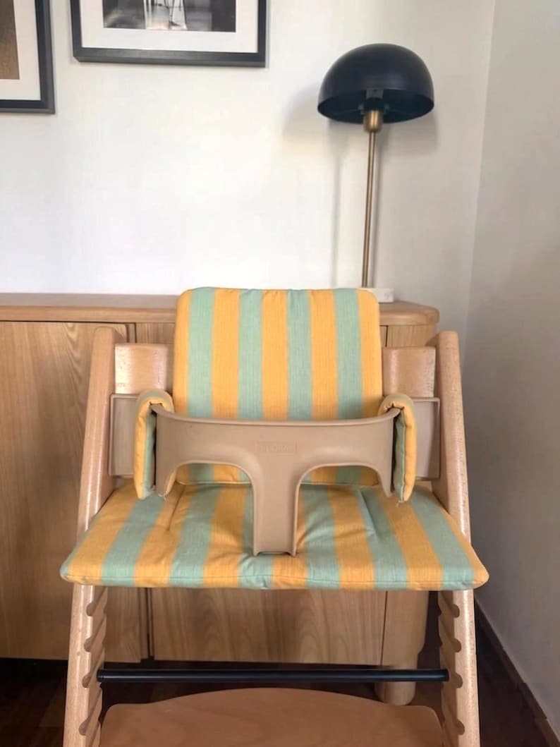Op de afbeelding: Houten kinderstoel met een beige veiligheidsbeugel en een zitkussen met gele en lichtgroene strepen. De stoel is ontworpen voor een kind en staat in een kamer met een lamp en houten meubels.