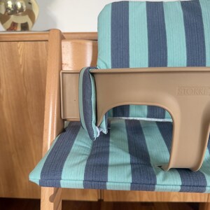 May include: A wooden high chair featuring a light brown plastic baby seat with the brand name STOKKE embossed on the front. The chair includes a padded seat and backrest cushion with a vertical striped pattern in teal and dark grey colours.