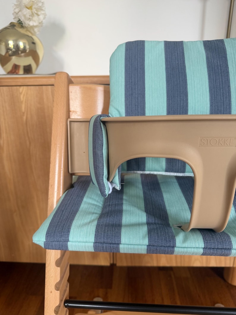 May include: A wooden high chair featuring a light brown plastic baby seat with the brand name STOKKE embossed on the front. The chair includes a padded seat and backrest cushion with a vertical striped pattern in teal and dark grey colours.